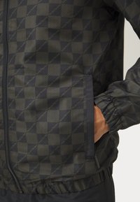 Giacca leggera in colori scuri, caratterizzata da un motivo geometrico a quadrati. Include polsini elasticizzati e una tasca con zip per maggiore funzionalità.