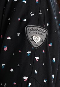Tissu noir avec de petits motifs en forme de goutte blancs, roses et bleus, avec un patch triangulaire de la marque Ragwear avec un emblème en forme de cœur.