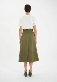 Jupe verte olive, longueur genoux, avec une taille ajustée et une silhouette en A. Associée à un t-shirt blanc et des chaussures à talons blocs vertes.