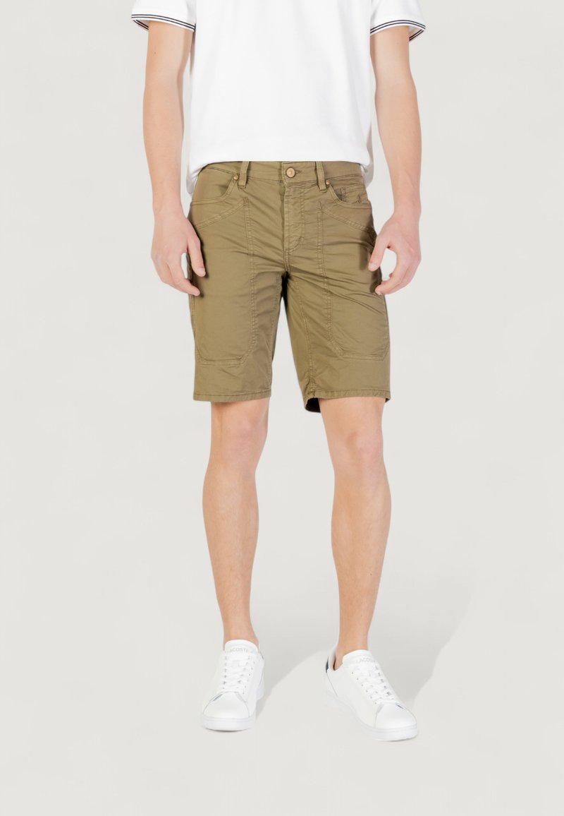 Pantaloni cargo verde oliva con una vestibilità rilassata, caratterizzati da molteplici tasche e dettagli di cucitura. Indossati con una camicia bianca a maniche corte e sneakers bianche.