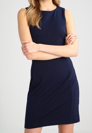 Femme portant une robe bleu marine sans manches, longueur genou, debout les bras croisés devant un fond clair uni.