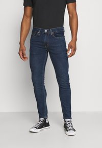 Jeans de mezclilla azul oscuro con un corte ajustado, que cuentan con cinco bolsillos y un cierre de botón metálico. Combinados con zapatillas de deporte negras de caña alta.