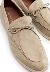 Mocassini in suede beige con punta arrotondata, lacci in pelle grezza e suola in gomma color crema. Presentano dettagli di cucitura accentuata.