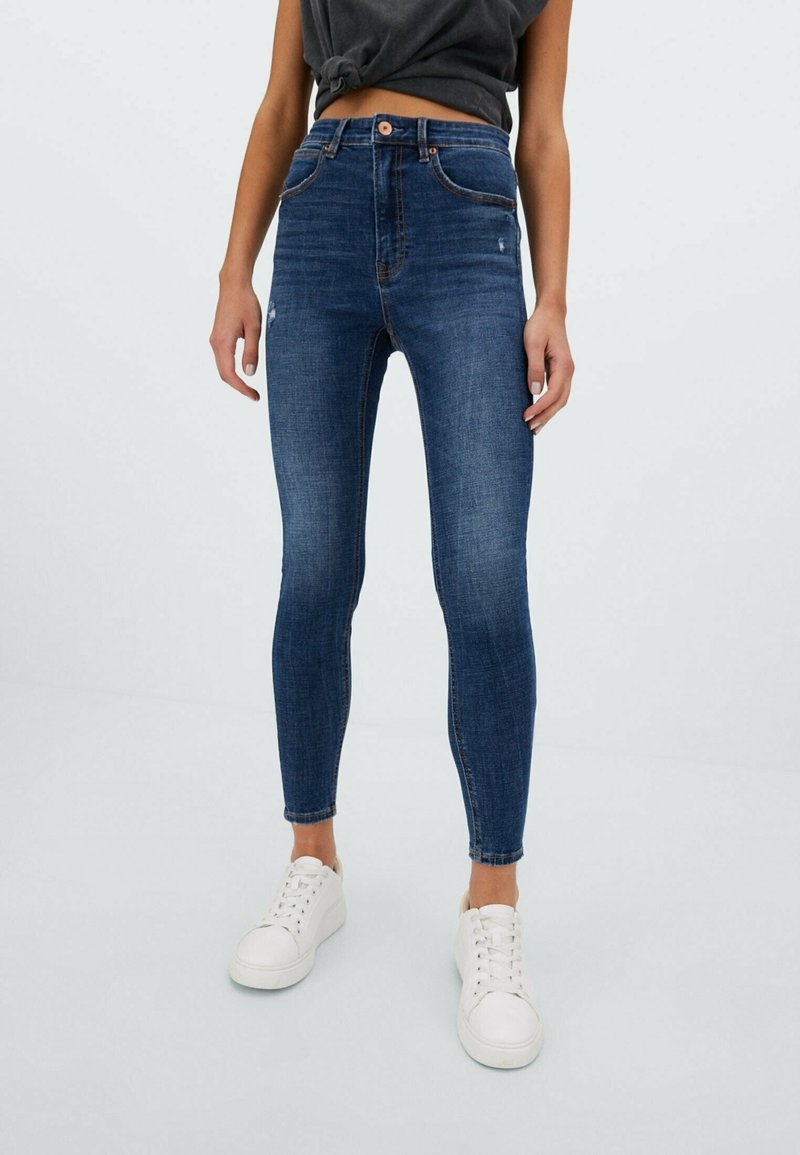 High-rise blue denim skinny jeans with slight fading, five-pocket design, and a zip-fly closure, paired with white sneakers.