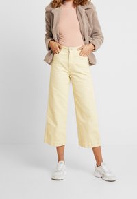 Gule brede bukser med høj talje, lavet af struktureret stof. Parret med en beige fuzzy jakke og hvide sneakers.
