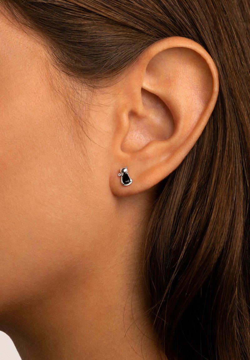 Pendientes pequeños de plata con una gema negra y un acento redondo, situados contra la oreja de un modelo y un fondo de cabello castaño.