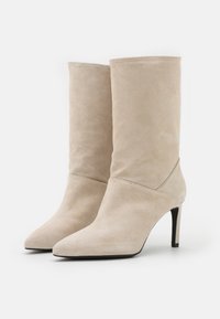Botas de tobillo de ante beige con punta afilada, textura suave, caña ajustada y un tacón alto y delgado. Diseño minimalista, sin herrajes visibles.