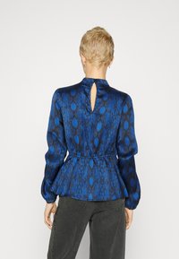 Blusa blu a fantasia con vita a peplo, maniche lunghe e dettaglio a chiave sul retro. Tessuto liscio con una leggera lucentezza. Abbinata a pantaloni scuri.