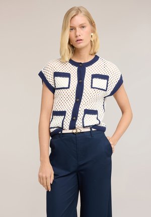 Mujer con cabello rubio que lleva un top de punto blanco sin mangas con ribete y botones azul marino, combinado con pantalones azul marino de talle alto y un cinturón.