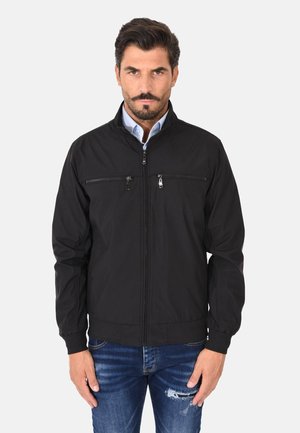 Uomo con giacca nera con cerniera sopra una camicia azzurra e jeans blu slavati, in piedi davanti a uno sfondo chiaro e uniforme.