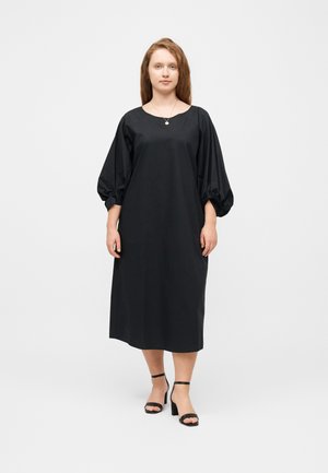 Femeie cu piele deschisă și păr șaten care poartă o rochie midi neagră lejeră cu mâneci bufante și sandale negre cu toc, stând pe un fundal alb.