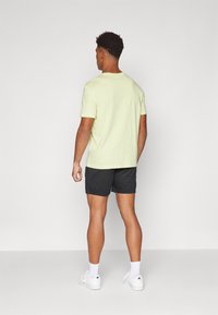 T-shirt amarelo claro com mangas curtas, combinada com calções pretos. Modelo a usar sapatilhas brancas, vista pela parte de trás contra um fundo simples.