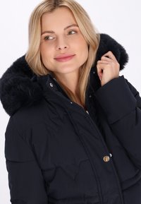 Marineblå pufferjakke med en faux fur-foring i hætten, lynlås foran og metalliske knapper. Diamantmønster quiltning er synlig.