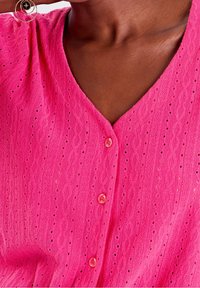 Cardigan en tricot rose avec un col en V, un motif texturé avec de petits trous et des boutons assortis. Matériau doux avec une sensation légère.
