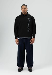 Zwarte oversized polo met een kraag en lange mouwen. Voorzien van witte tekst aan de voorkant. Gecombineerd met losse donkerblauwe jeans en zwarte schoenen.