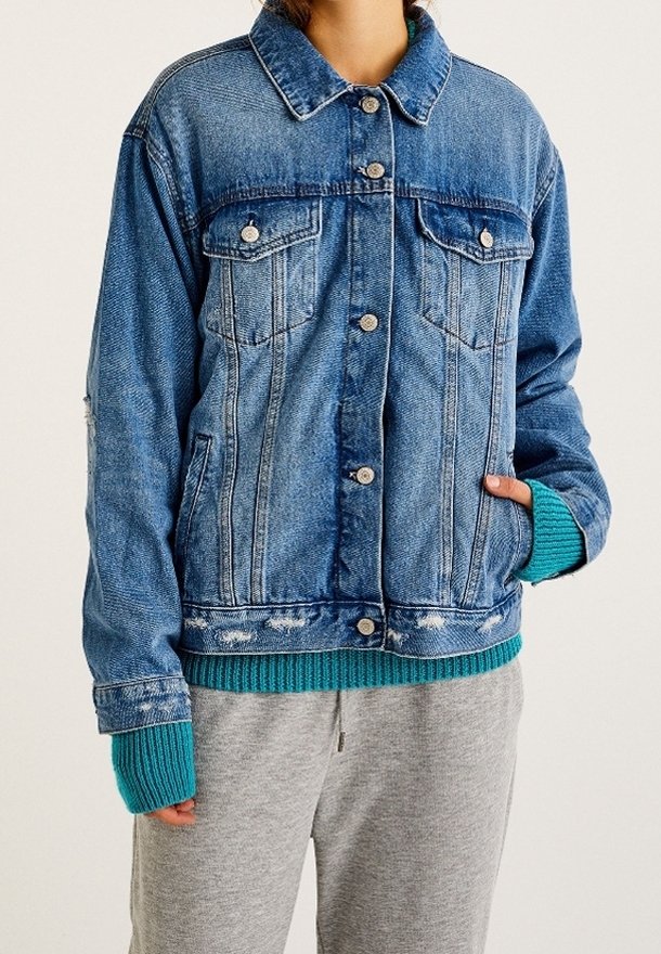 Personne portant une veste en jean bleue par-dessus un pull bleu sarcelle avec des poignets étendus, et un pantalon de survêtement gris clair, la main dans la poche de la veste.