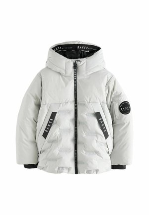 Jonge jongen met krullend blond haar, die een lichtgrijs gewatteerd jack met een hoge kraag en een zwarte broek draagt tegen een effen achtergrond.