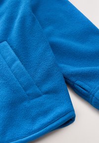 Jachetă din fleece albastru cu textură moale, având un buzunar frontal și manșete. Materialul este gros și cald, potrivit pentru vreme rece.