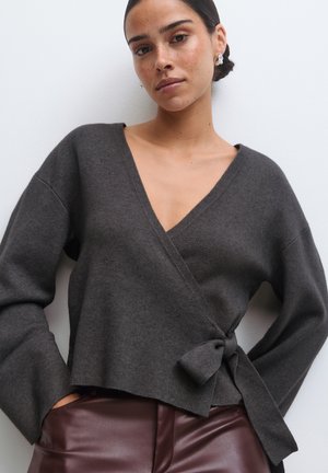 Vrouw met het haar in een lage knot draagt een donkergrijze overslagtrui vastgemaakt op de taille en bordeauxrode leren broek, staand tegen een effen achtergrond.