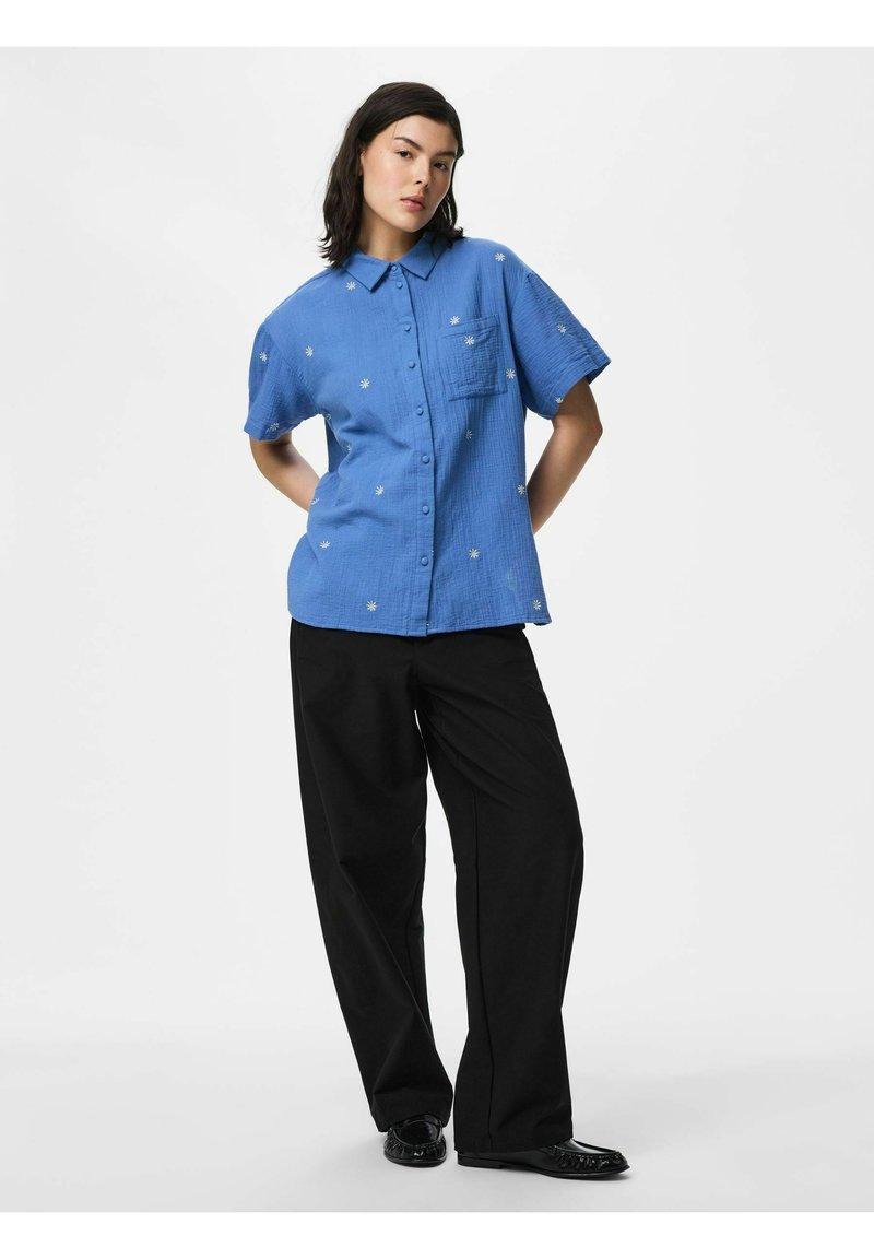 Femme portant une chemise bleue à manches courtes avec des imprimés de fleurs blanches, associée à un pantalon noir à jambes larges et des chaussures noires.