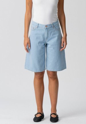 Személy világoskék térdig érő farmer rövidnadrágot, fehér ujjatlan felsőt, fekete lapos cipőt visel, és látható kar tetoválása van szürke háttér előtt.