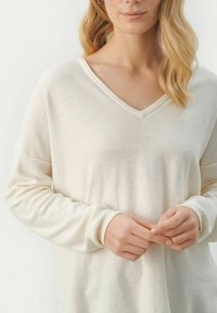 Cremefarbener V-Ausschnitt-Pullover aus weichem Material, mit langen Ärmeln und gerippten Bündchen. Die lockere Passform betont eine glatte Textur mit einem dezenten Schimmer.