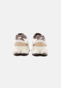 Brūnas un krēma krāsas New Balance sporta apavi no aizmugures, ar teksturētu virsējo daļu, mīkstu zoli, logotipa akcentiem un apaļo purngali.