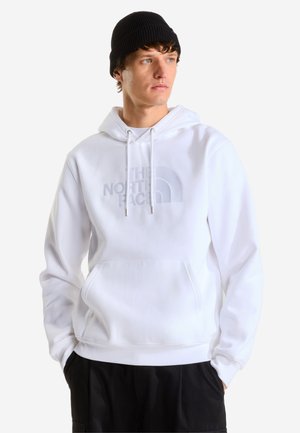 Ung mand iført en hvid The North Face hættetrøje og sort beanie, med hænderne i lommerne, kigger til siden på en ensfarvet baggrund.