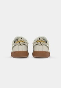 Biele kožené tenisky s textúrovaným povrchom, hnedá gumová podrážka a leopardí vzor na päte s nápisom "Reebok" v zlatej farbe.