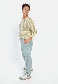Gestreepte lange mouwen top in lichtgeel en groen. Lichtblauwe ribfluweel broek met zijzakken. Witte sneakers met grijze accenten.