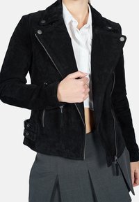 Veste moto en daim noir avec une fermeture éclair asymétrique, des boutons-pression et des poches latérales zippées, présentant une silhouette ajustée et une texture douce.