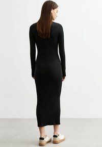 Robe noire à manches longues en tissu extensible, au design moulant, longueur cheville, associée à des chaussures légères avec un talon contrastant.