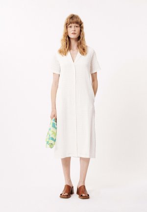 Weißes kurzärmliges Kleid mit V-Ausschnitt, knielang, aus strukturiertem Stoff und mit Knopfdetails; Modell hält ein gemustertes grün-blaues Handtuch.
