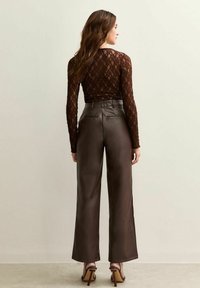 Een vrouw gezien van achteren, gekleed in een doorschijnende bruine kant blouse met lange mouwen, hoogwaist wijdvallende bruine leren broek en bruine hoge hakken.