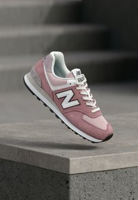Baskets New Balance 574 roses et blanches avec des lacets blancs, flottant au-dessus d'un rebord en béton avec des marches floues en arrière-plan.