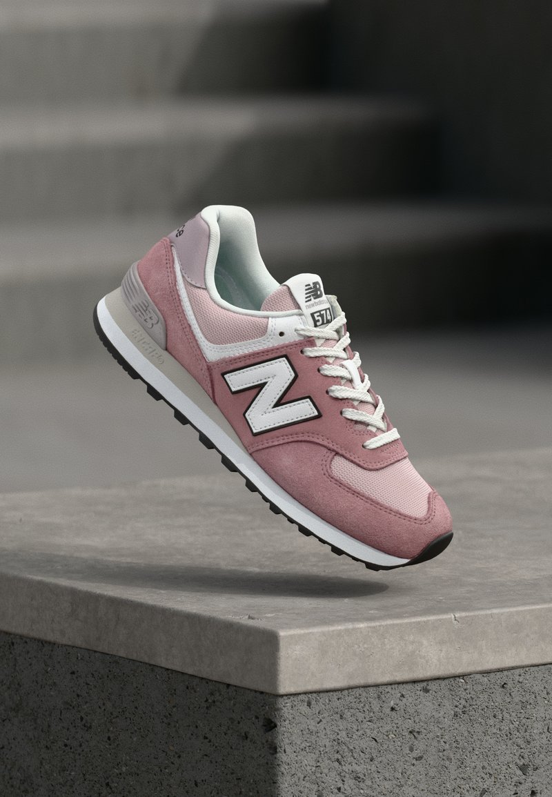 Baskets New Balance 574 roses et blanches avec des lacets blancs, flottant au-dessus d'un rebord en béton avec des marches floues en arrière-plan.