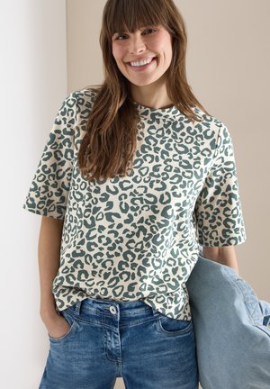 Femme souriante aux longs cheveux bruns portant un haut à manches courtes imprimé léopard vert et blanc, un jean bleu, et tenant une veste bleu clair.