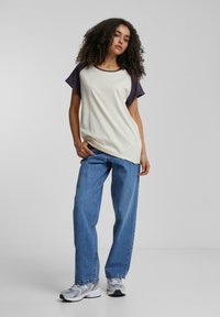 Kortærmet, cremet T-shirt med lilla raglanærmer, parret med lyseblå lige ben jeans og grå sneakers, sat mod en neutral baggrund.