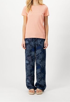 Kobieta ubrana w brzoskwiniowy t-shirt z krótkim rękawem, granatowe szerokie spodnie z wzorem liści palmowych oraz plecione sandały na platformie.
