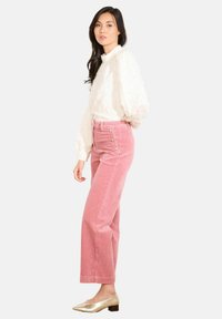 Žena oblečená v bielej štruktúrovanej blúzke, nohaviciach z koridury s vysokým pásom v ružovej farbe so bočnými gombíkmi a zlatých topánkach na nízkom podpätku na bielom pozadí.