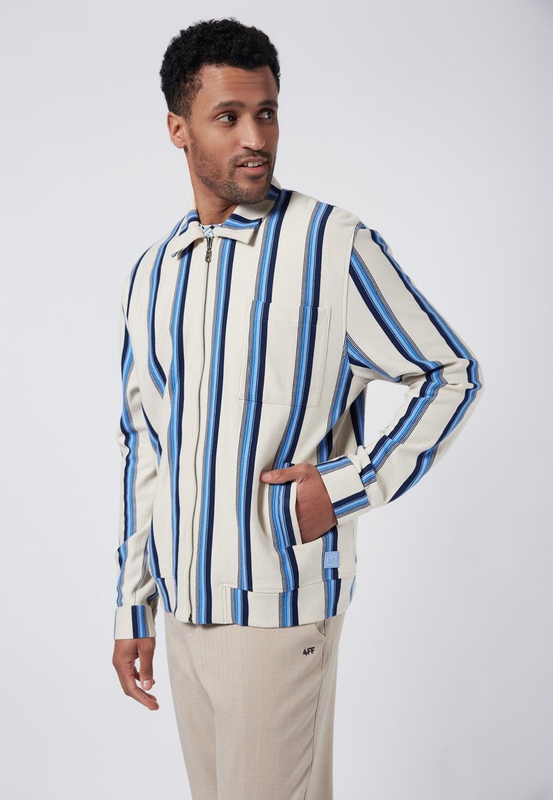 Randig jacka i krämfärgad med blå och navy ränder, med dragkedjestängning, krage och bröstficka. Kombinerad med ljusbeiga byxor.