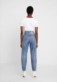 Person som står med ryggen mot kameran, iklädd en vit kortärmad skjorta, höga blå jeans med logotypmärke och vita klackiga ankelstövlar.