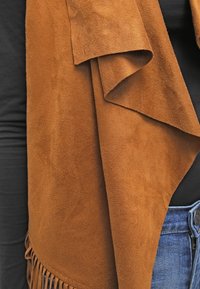 Chal de ante sintético marrón con diseño drapeado y detalle de flecos en el borde inferior, colocado sobre una camiseta negra.