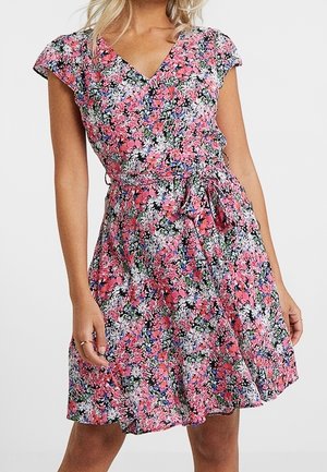 Robe portefeuille fleurie avec des fleurs roses, blanches et bleues, manches courtes, col en V et taille nouée, portée par une femme.