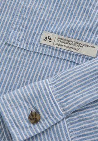 Ljusblå och vit randig tygskjorta med knäppbar manschett och en etikett som läser "Underground Denim Association of Nineteen Eighty Two."