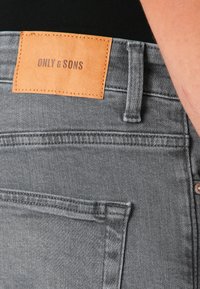 Jeans de mezclilla gris con una etiqueta de cuero que dice "ONLY & SONS". Incluye bolsillos traseros y detalles de costura en contraste.