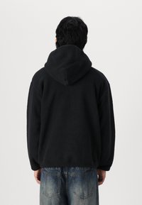 Černá fleecová mikina s velkou kapucí a strukturovaným materiálem, spárovaná se světlými modrými džínami s dírami. Žádné viditelné logo ani ozdoby.