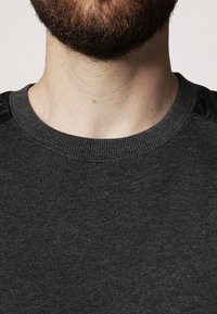 Suéter gris oscuro con cuello acanalado y detalles de tela negra texturada en los hombros. Material de punto suave con un diseño clásico de cuello redondo.