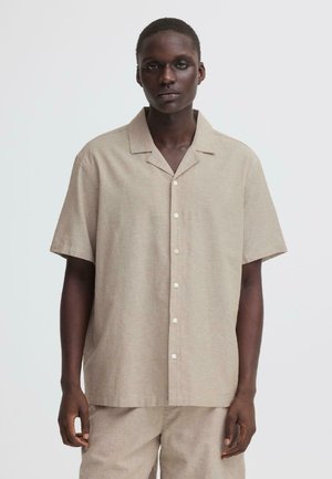 Mężczyzna w beżowej koszuli na krótki rękaw z zapięciem na guziki oraz pasujących spodniach, stojący na tle jasnego, jednolitego tła.