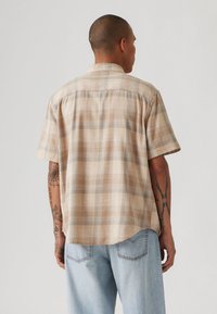 Short-sleeved, beige plaid shirt with a relaxed fit, featuring light gray accents and a curved hem. Woven texture is visible.